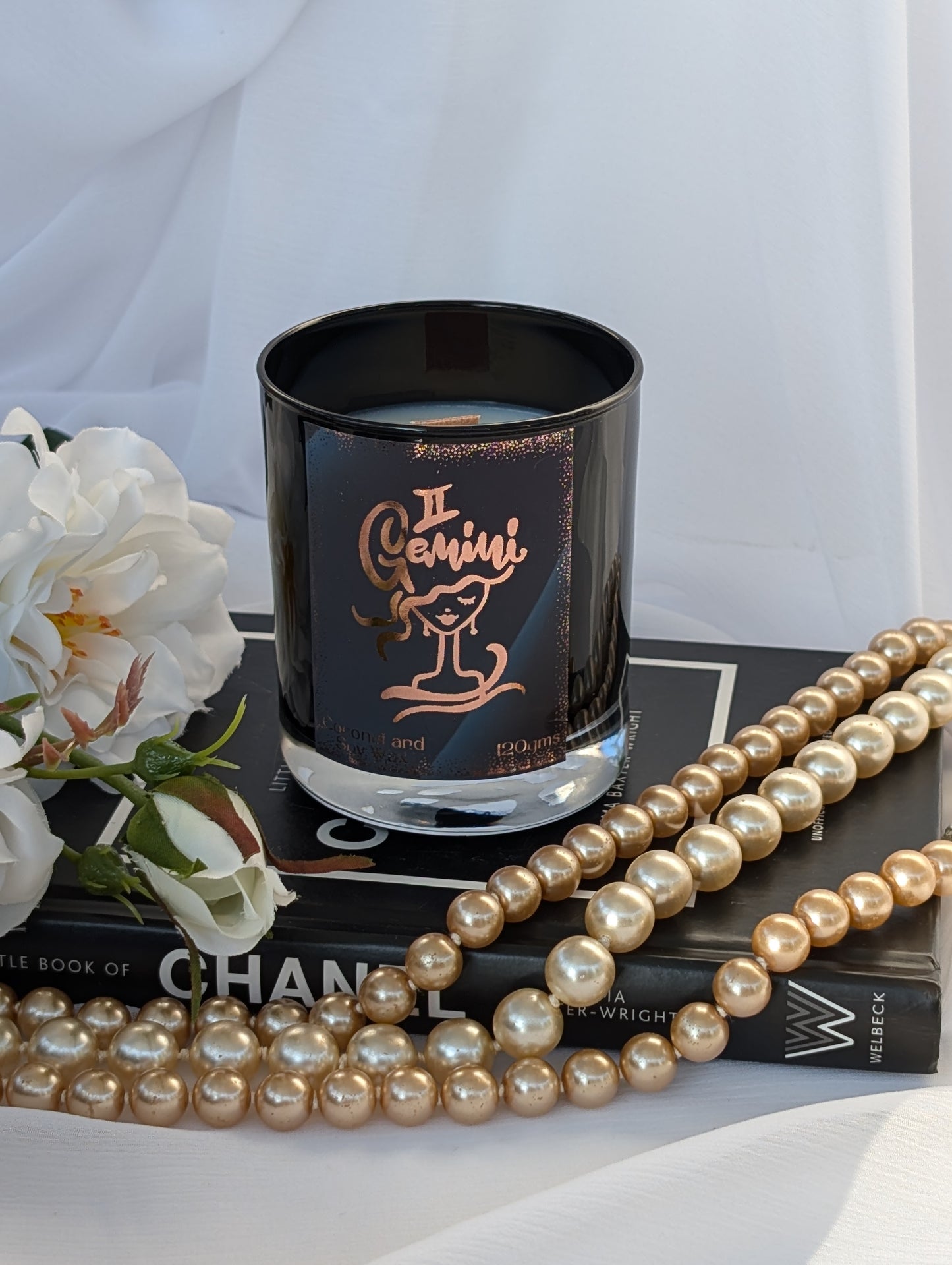 A black glass candle jar with a rose-gold Gemini zodiac design, styled on a black book with white flowers and pearl strands in the foreground against a white draped fabric background.