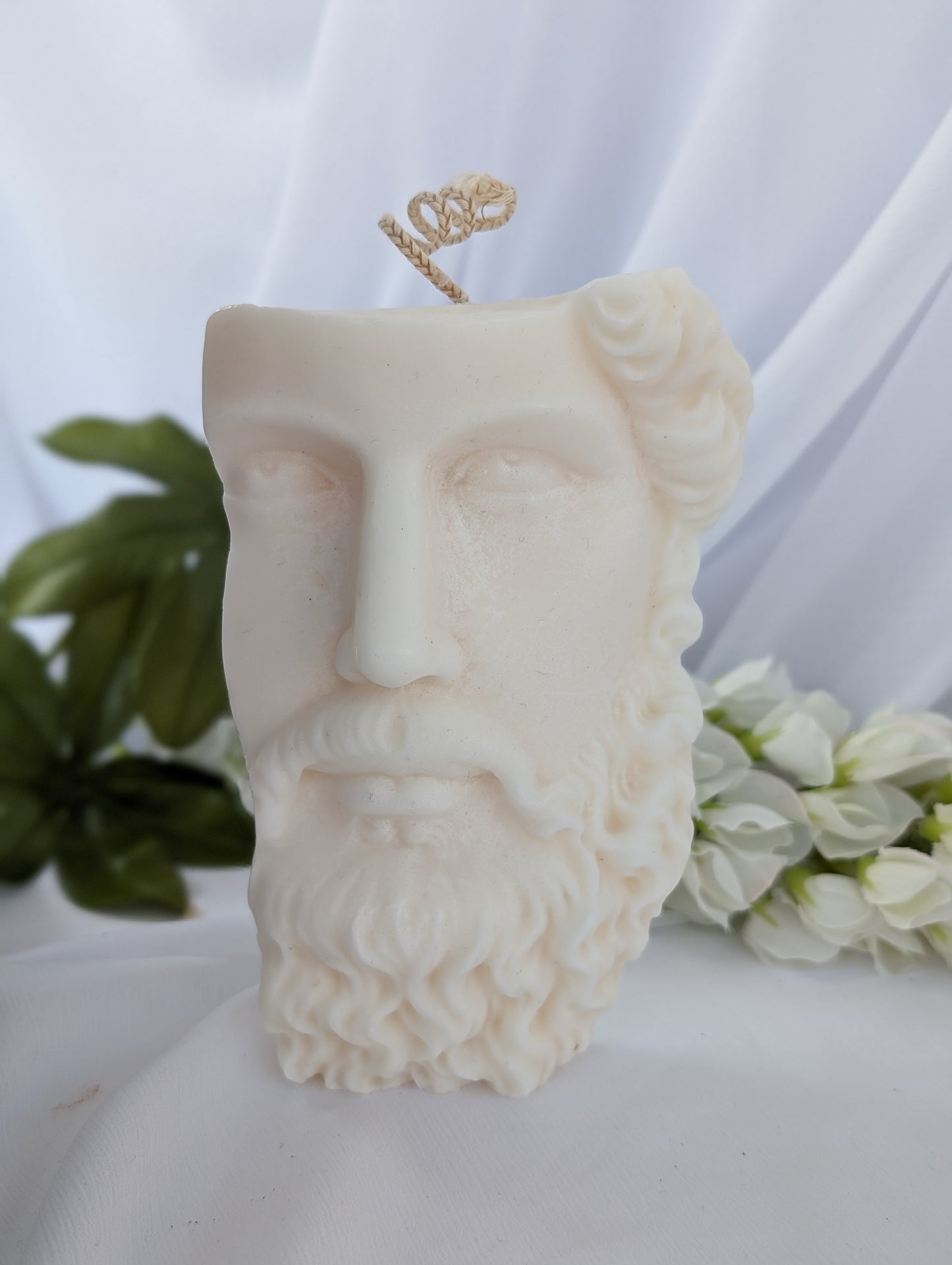 Candle shaped like a classical Greek bust on a white background with flowers in the background