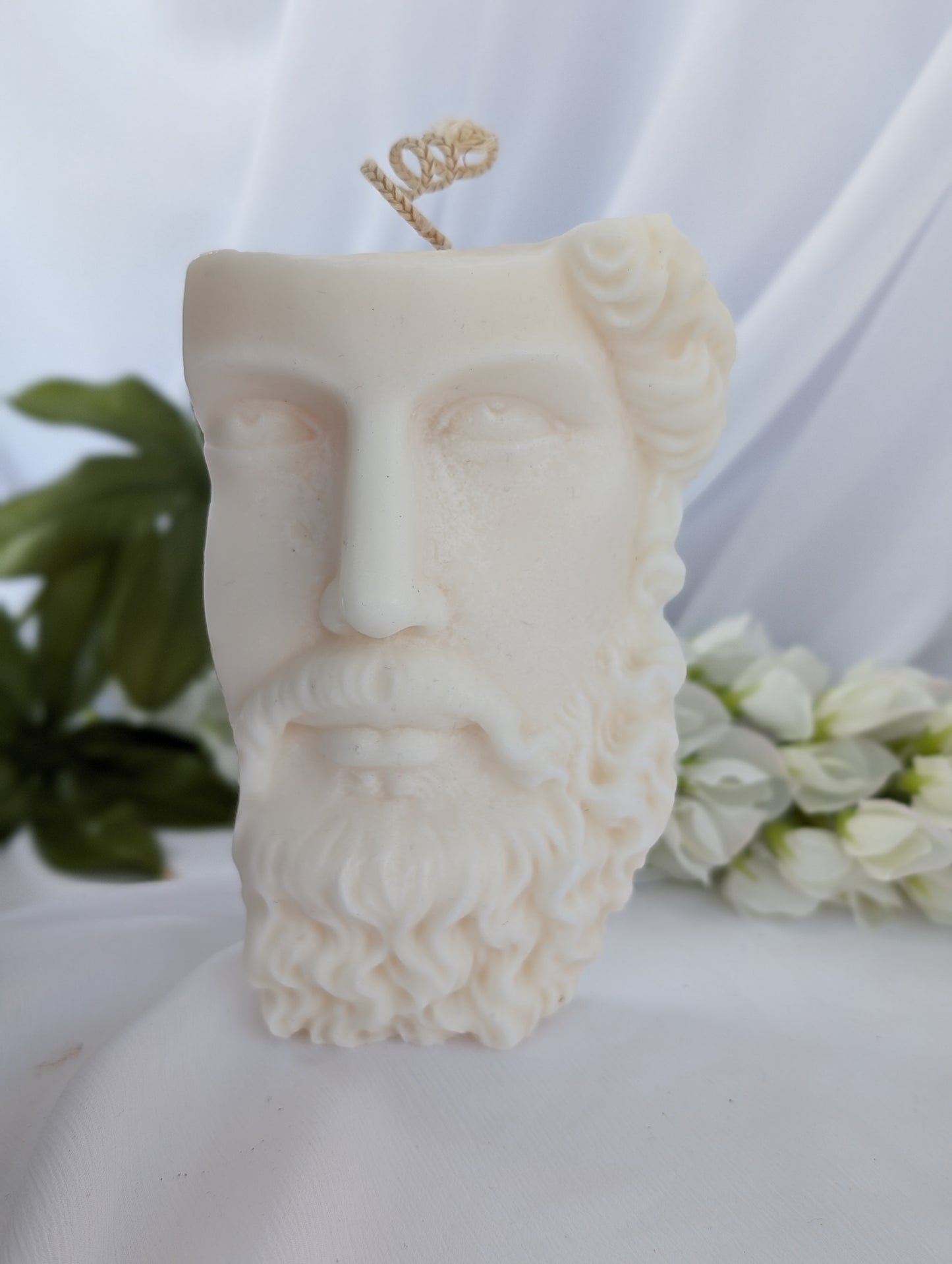 Candle shaped like a face with a beard on a white surface with greenery in the background