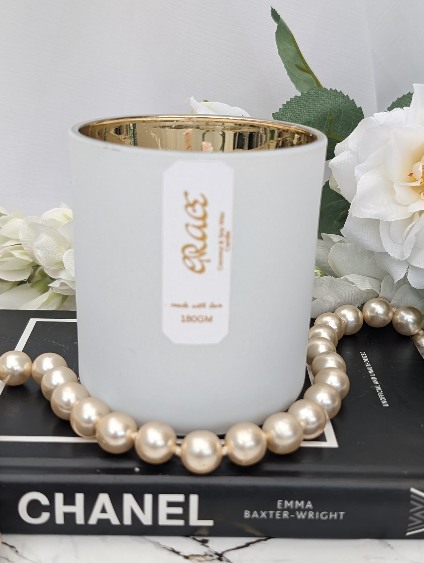 Luxury white candle with gold interior on a Chanel book, surrounded by pearls and white flowers.