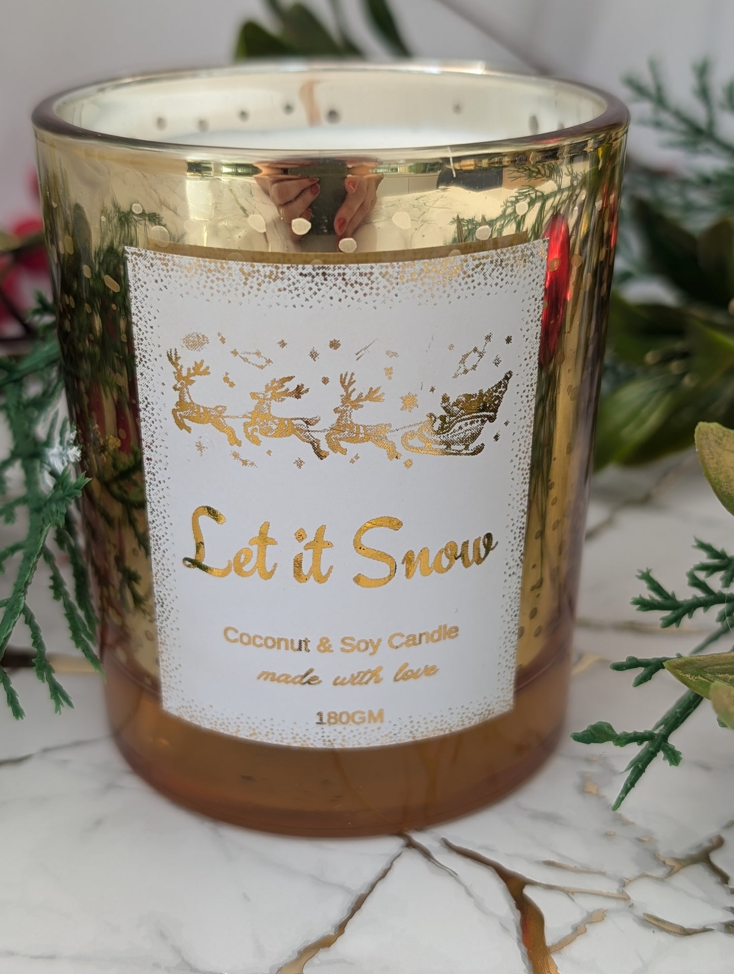 Candle with 'Let it Snow' label and reindeer design, surrounded by greenery on a marble surface.