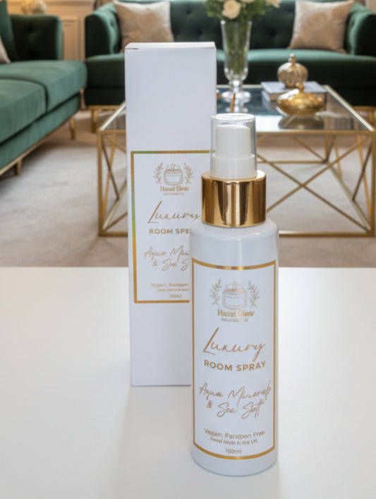 A white bottle of 'Luxury Room Spray' with a gold collar and clear spray nozzle, standing in front of its matching white packaging box on a neutral gray surface.