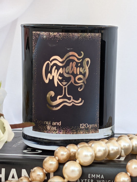 Black and gold Aquarius zodiac candle - black label featuring metallic rose gold foil script  The candle is styled on a book with a gold pearl necklace draped over it and white flowers at the side.