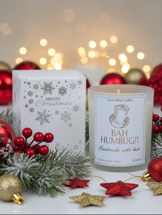 A frosted glass jar candle labeled 'BAH HUMBUG! Handmade with love' by Hozed Glow Candles, shown next to its white Christmas-themed gift box with silver snowflakes and 'Merry Christmas' text, set against a background of red poinsettias.