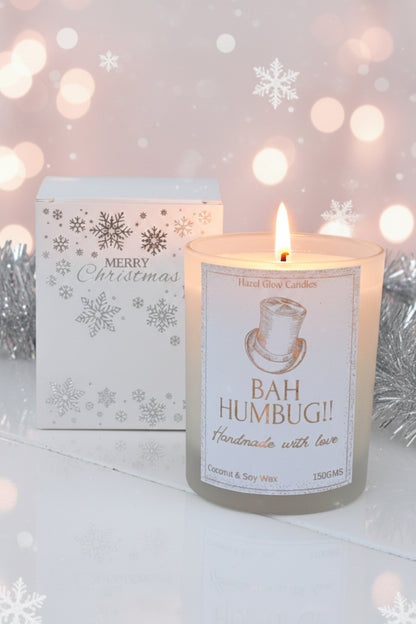A frosted glass jar candle labeled 'BAH HUMBUG!!' by Hexed Glow Candles, shown next to its white Christmas-themed gift box with silver snowflakes and 'Merry Christmas' text, against background of silver tinsel, snowflakes and bokeh