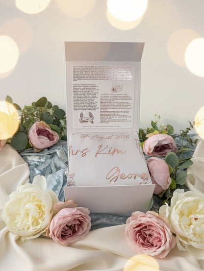 A white keepsake box shown open, with printed instructions on the inside lid and a white fabric insert featuring rose-gold foil script reading 'Mrs Kim' and 'Georg' with '08th August 2026' above. Pink flowers and blue satin fabric surround the box.