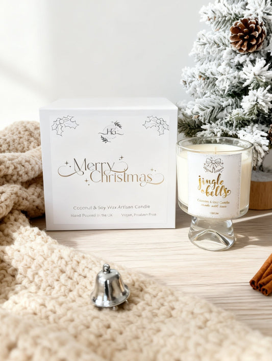 A festive Christmas candle set on a light wood surface, featuring a white box labeled 'Merry Christmas' and a glass jar candle with gold lettering, styled with a cream knit blanket, cinnamon sticks, a small silver bell, and a flocked Christmas tree in the background.