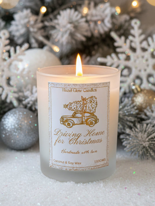 A frosted glass candle jar with a white and gold label reading 'Hazel Glow Candles' and 'Driving Home for Christmas,' featuring an illustration of a vintage car carrying a Christmas tree. The candle is photographed on a white surface with a softly blurred Christmas background.