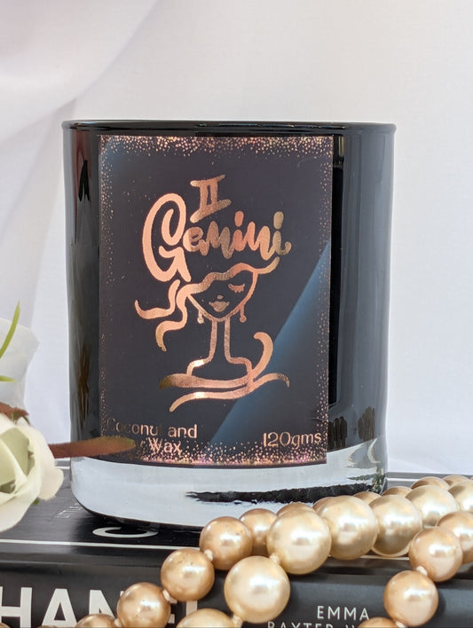 A Gemini-themed coconut and wax candle in a black glass jar with a metallic rose gold label, styled on a book with pearl beads in the foreground.