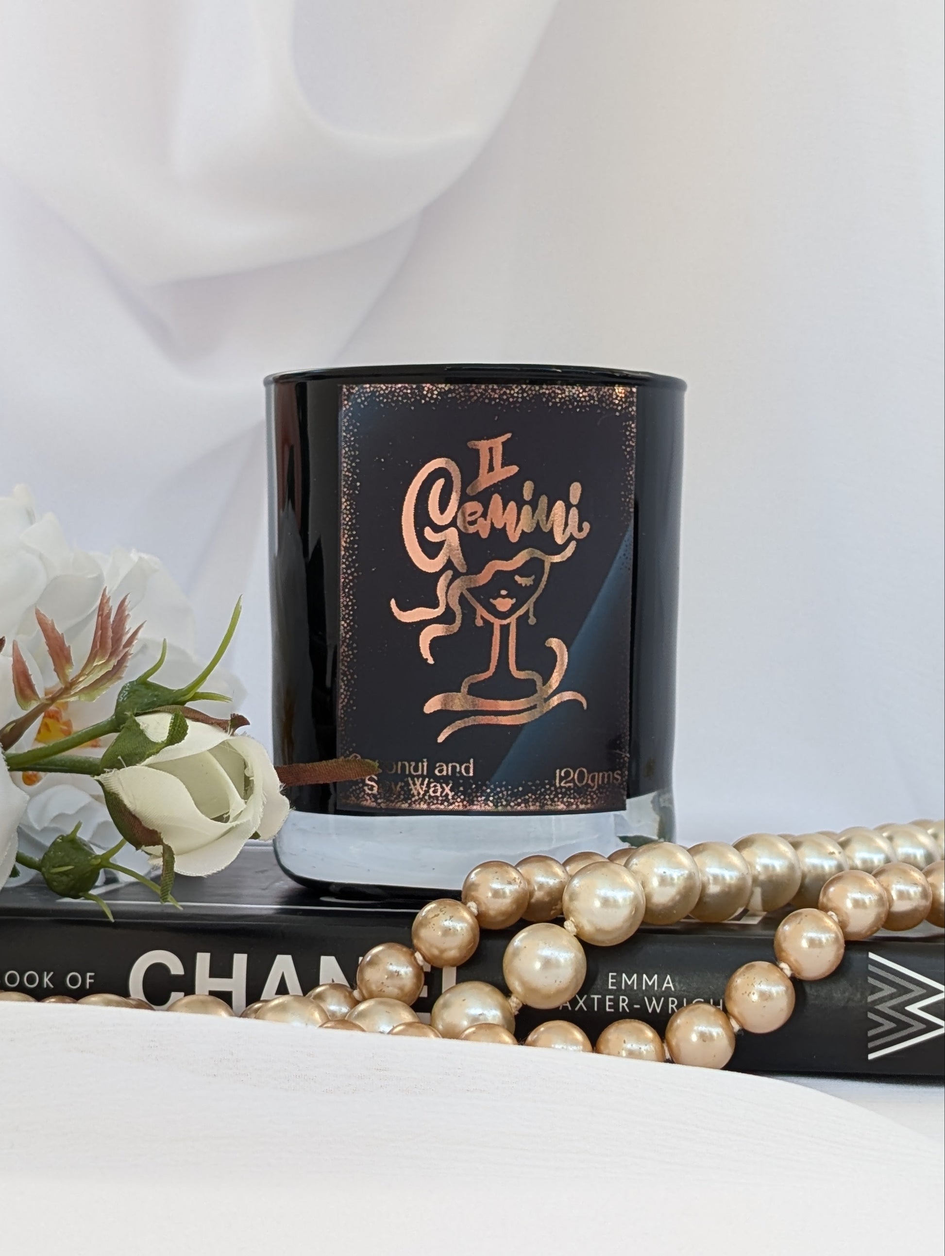 A black glass candle jar with a metallic rose gold Gemini zodiac design, styled on a book with white flowers and a pearl necklace.