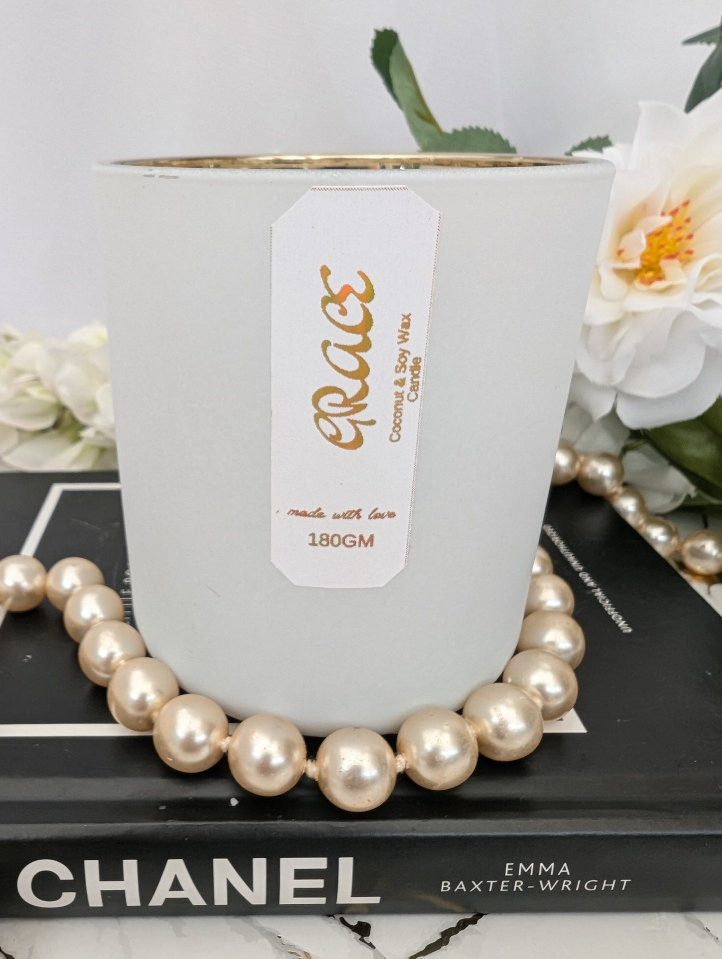 Luxury White candle with a gold foiled label on a Chanel book, surrounded by pearls and flowers.