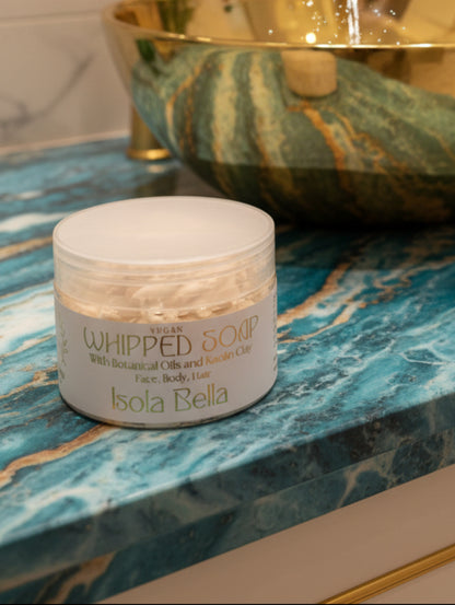 A jar of Isola Bella Vegan Whipped Soap with botanical oils and kaolin clay, featuring a white label with gold lettering and a clear lid, set on a gray marble surface