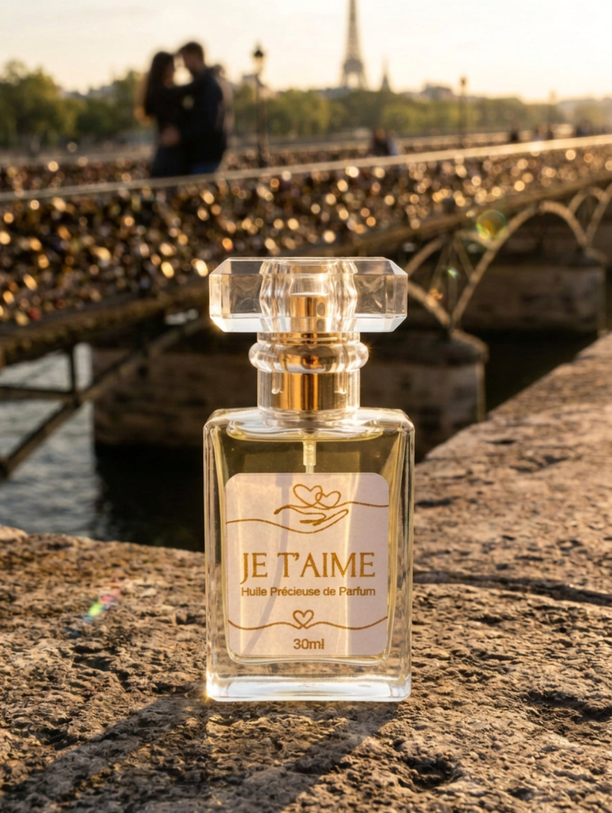 A 30ml bottle of 'Je T'Aime' precious perfume oil with a clear glass body, gold spray pump, and a white label with gold lettering, set against a Parisian background and the famous bridge of padlocks