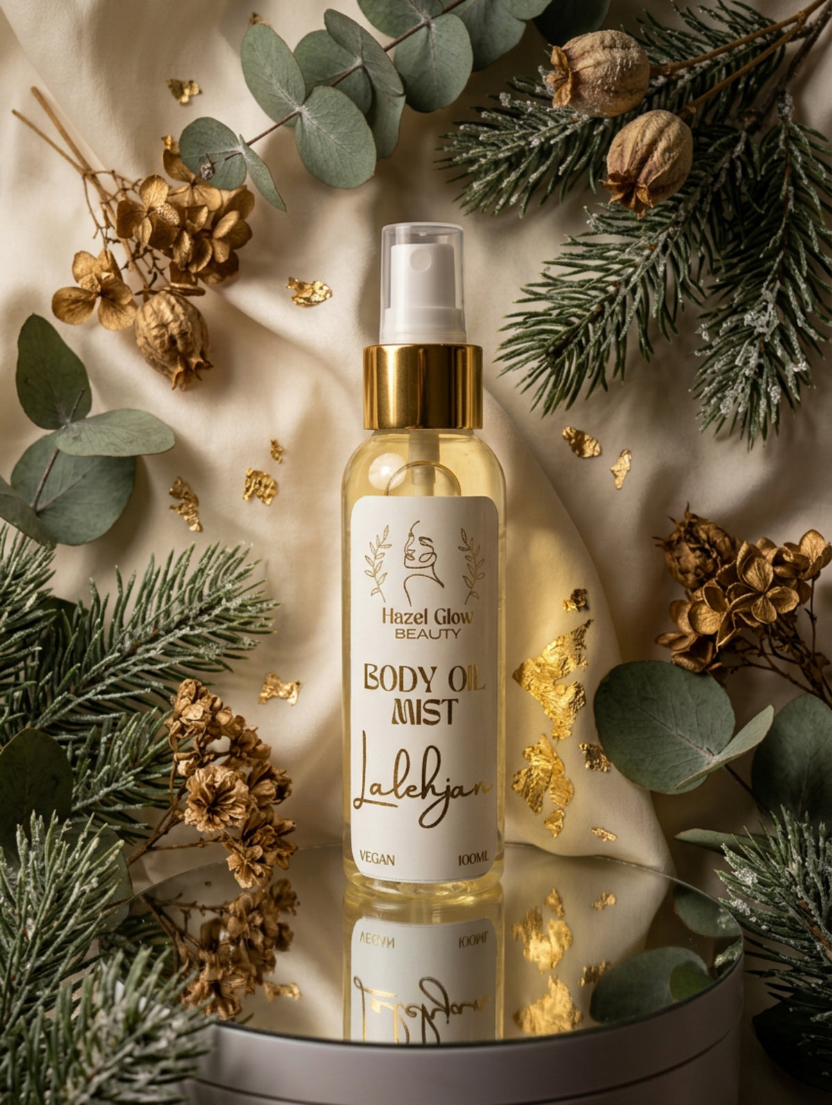 A bottle of Hazel Glow Beauty Body Oil Mist with a white label and gold collar, displayed on a reflective silver tray with sage green Christmas foliage in the background.