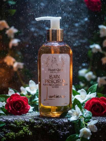 A 200ml bottle of Hazel Glow Beauty Bain Precisieux Fine Indulgence Body Wash, featuring a white pump dispenser, a gold collar, and a white label with rose gold text, set against a background of flowers and falling water drops.