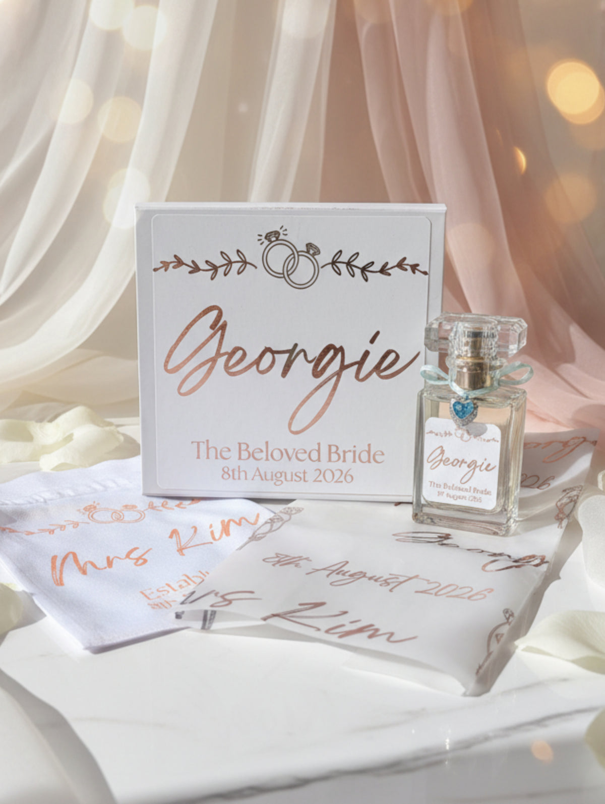 Personalized wedding gift set including a small clear perfume bottle with silver charm, keepsake box with rose gold script reading 'Georgie', satin drrawstring bag and personalised foiled vellum, against a luxury bridal background