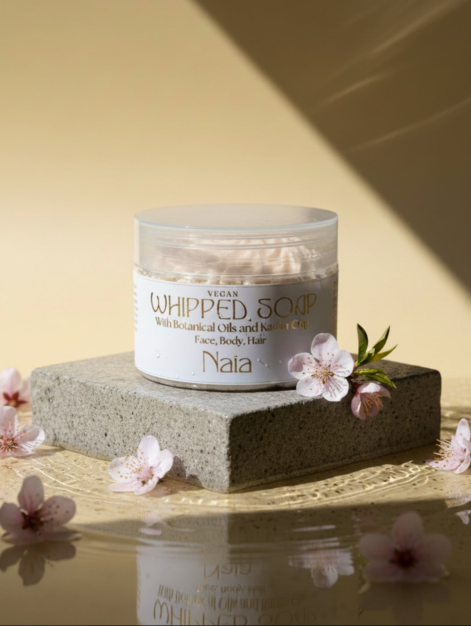 A jar of Naia Vegan Whipped Soap with botanical oils and kaolin clay, featuring a white label with gold lettering on a clear plastic container, set on a rough stone in water with scatter peach blossom