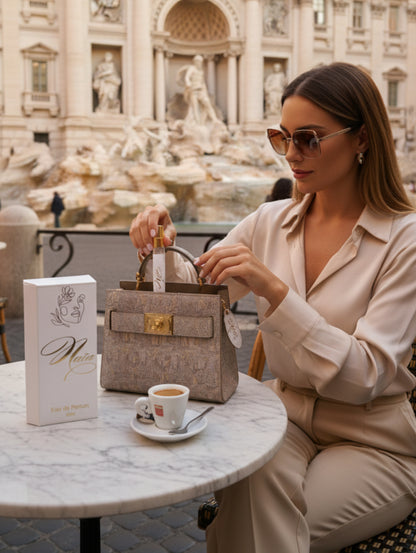A stylish woman at a cafe in front of the Trevi fountain taking Naia perfume by Hazel Glow out of her bag.  Naia box is on the table in front of her
