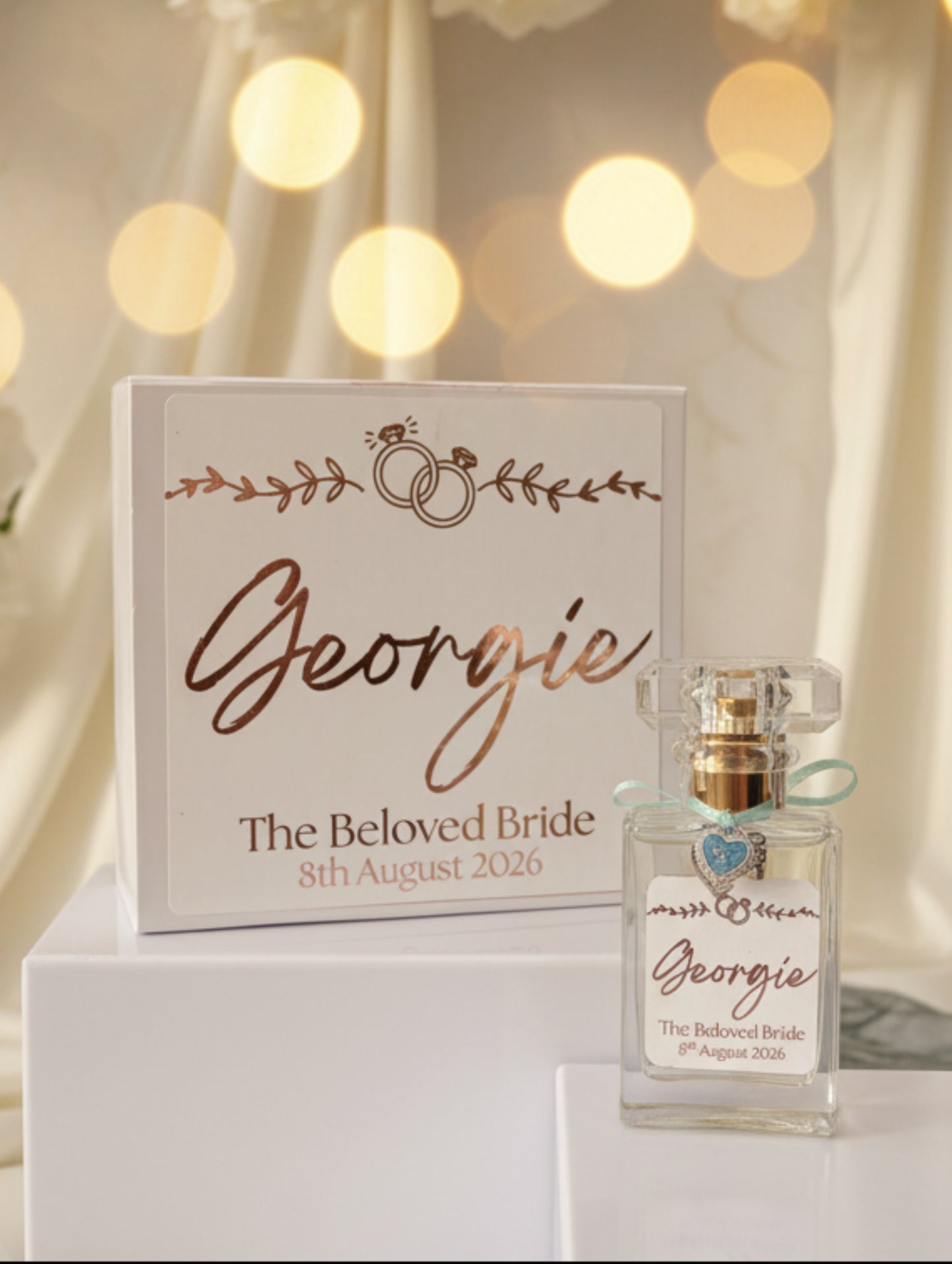 A personalized perfume bottle with a clear glass body, gold cap, and light blue ribbon, displayed next to its white presentation box. Both pieces feature custom rose gold script reading 'Georgie' and 'The Beloved Bride' with the date '8th August 2026' on a neutral background.