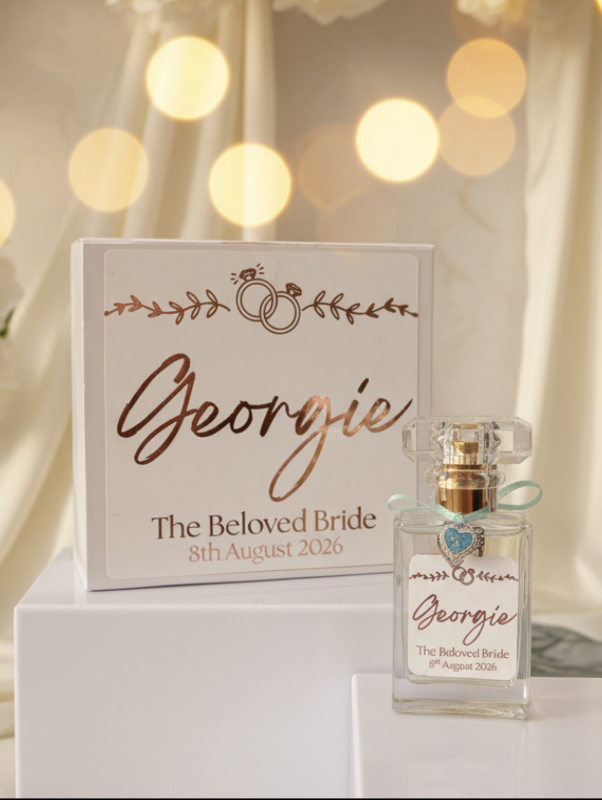 A personalized perfume bottle with a clear glass body, gold cap, and light blue ribbon, displayed next to its white presentation box. Both pieces feature custom rose gold script reading 'Georgie' and 'The Beloved Bride' with the date '8th August 2026' on a neutral background.