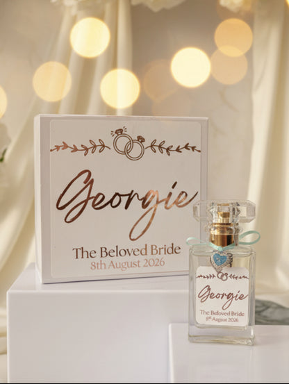 A personalized perfume bottle with a clear glass body, gold cap, and light blue ribbon, displayed next to its white presentation box. Both pieces feature custom rose gold script reading 'Georgie' and 'The Beloved Bride' with the date '8th August 2026' on a neutral background.