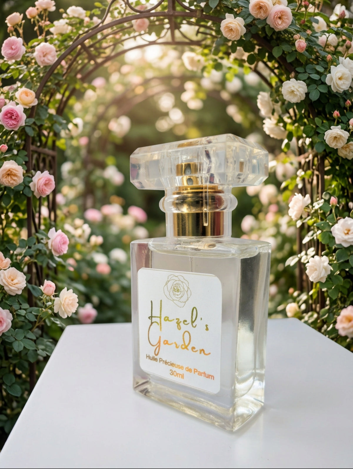 Hazel's Garden precious perfume oil.  In a clear glass bottle with faceted lid, white label with gold foil detail shown on a white table with flowers in the background