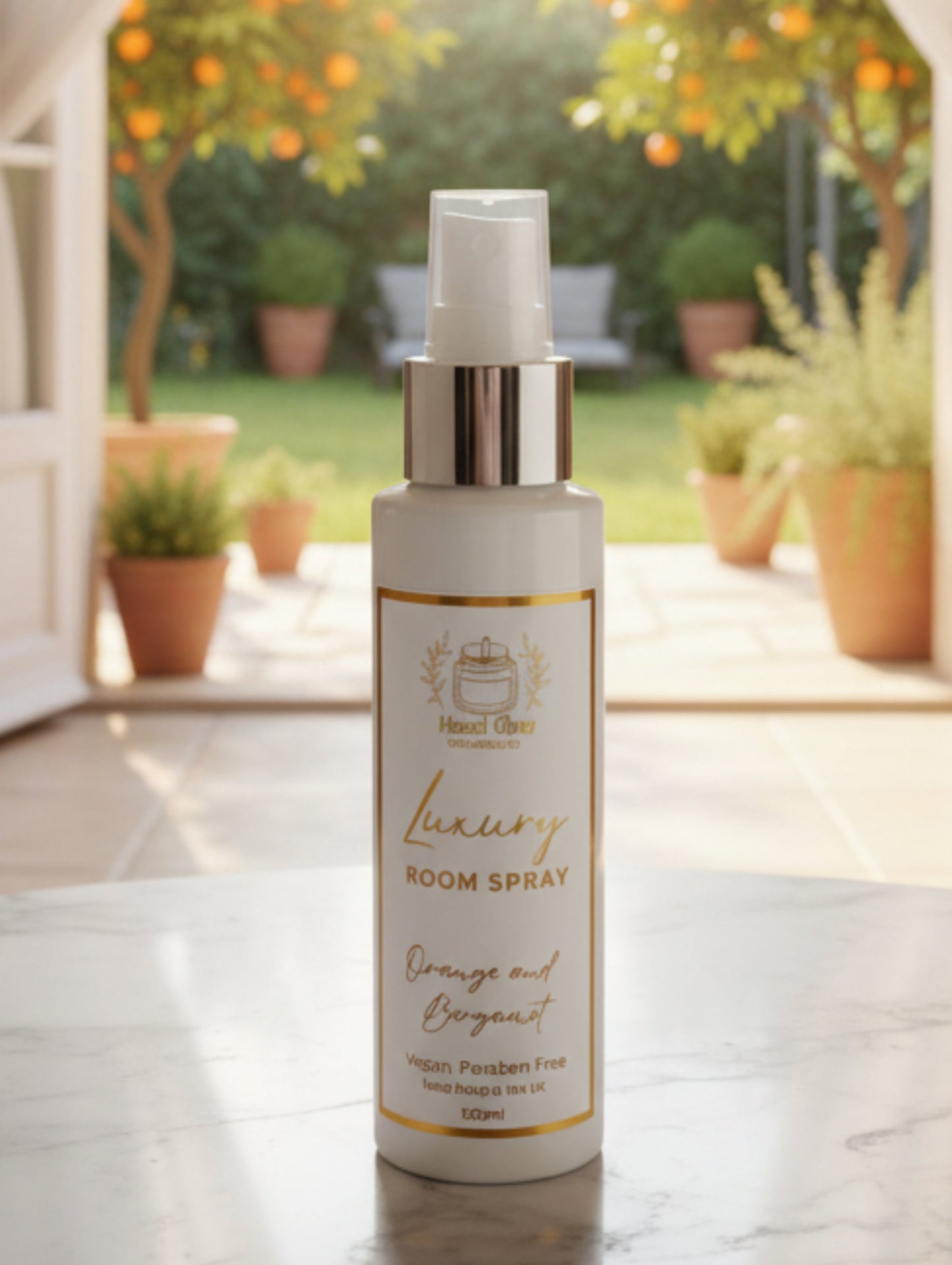 For your home fragrance rituals - A 100ml white bottle of  Luxury Room Spray with a silver pump and a white label featuring gold foiled text, set on a marble table with orange trees in the background