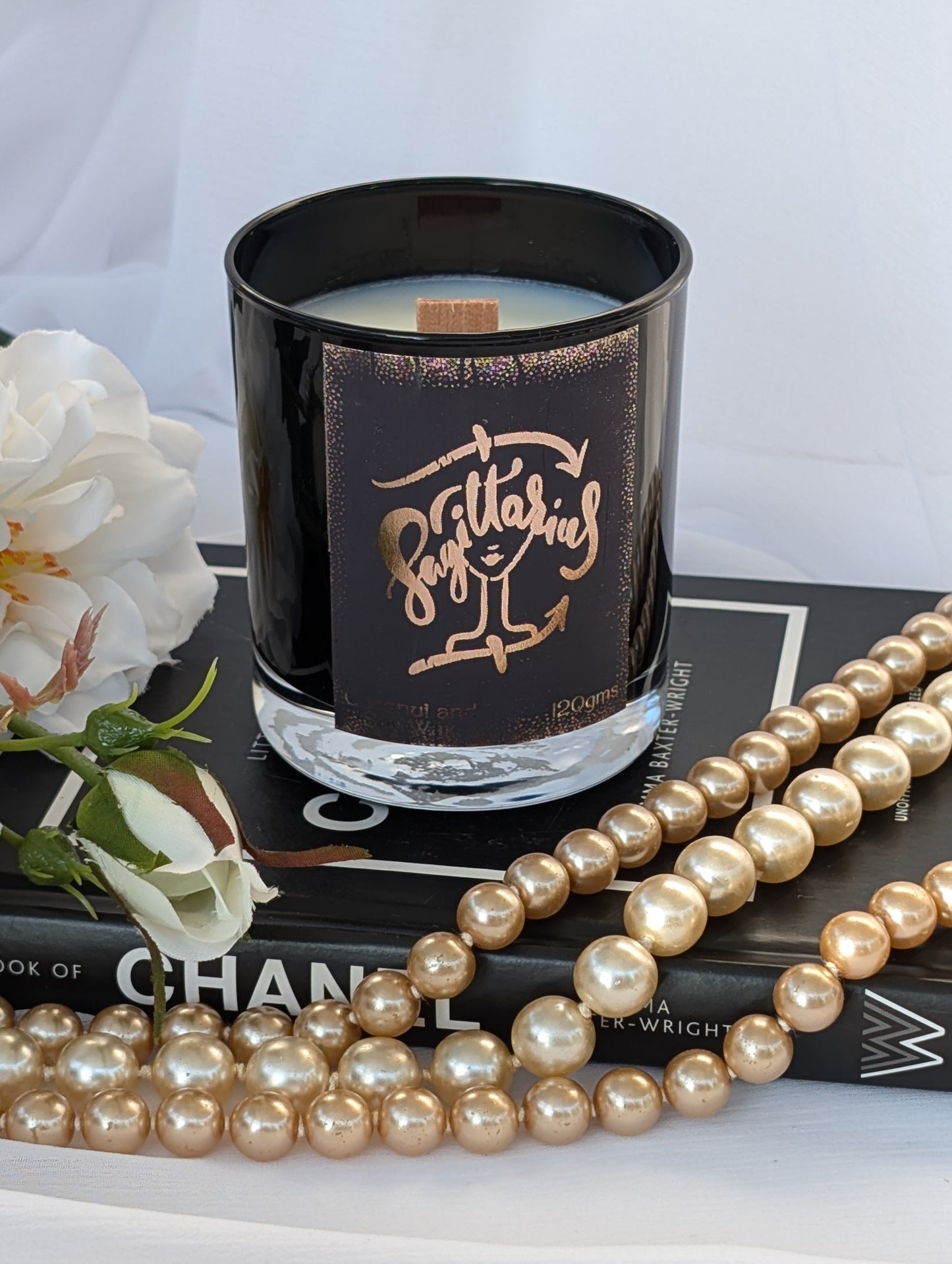 A black glass candle jar with a metallic rose-gold label Sagittarius sits on a black book, surrounded by white flowers and draped with a strand of faux pearls against a white fabric backdrop.