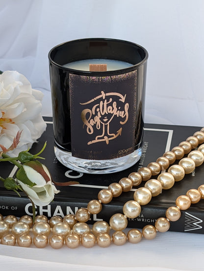 A black glass candle jar with a metallic rose-gold label Sagittarius sits on a black book, surrounded by white flowers and draped with a strand of faux pearls against a white fabric backdrop.