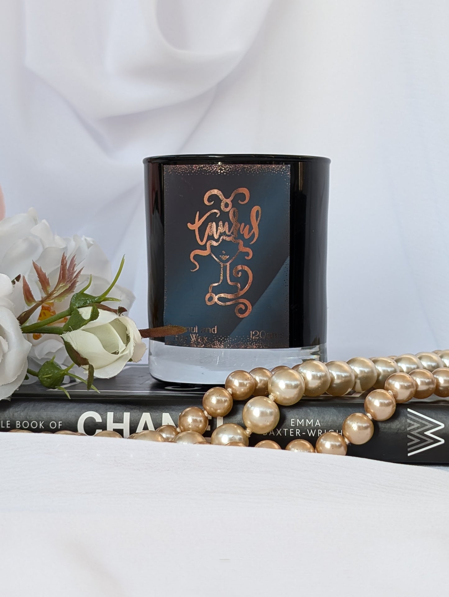 Black and Gold Taurus candle - sits on a book, styled with white flowers and a strand of gold pearls against a soft white draped fabric background.