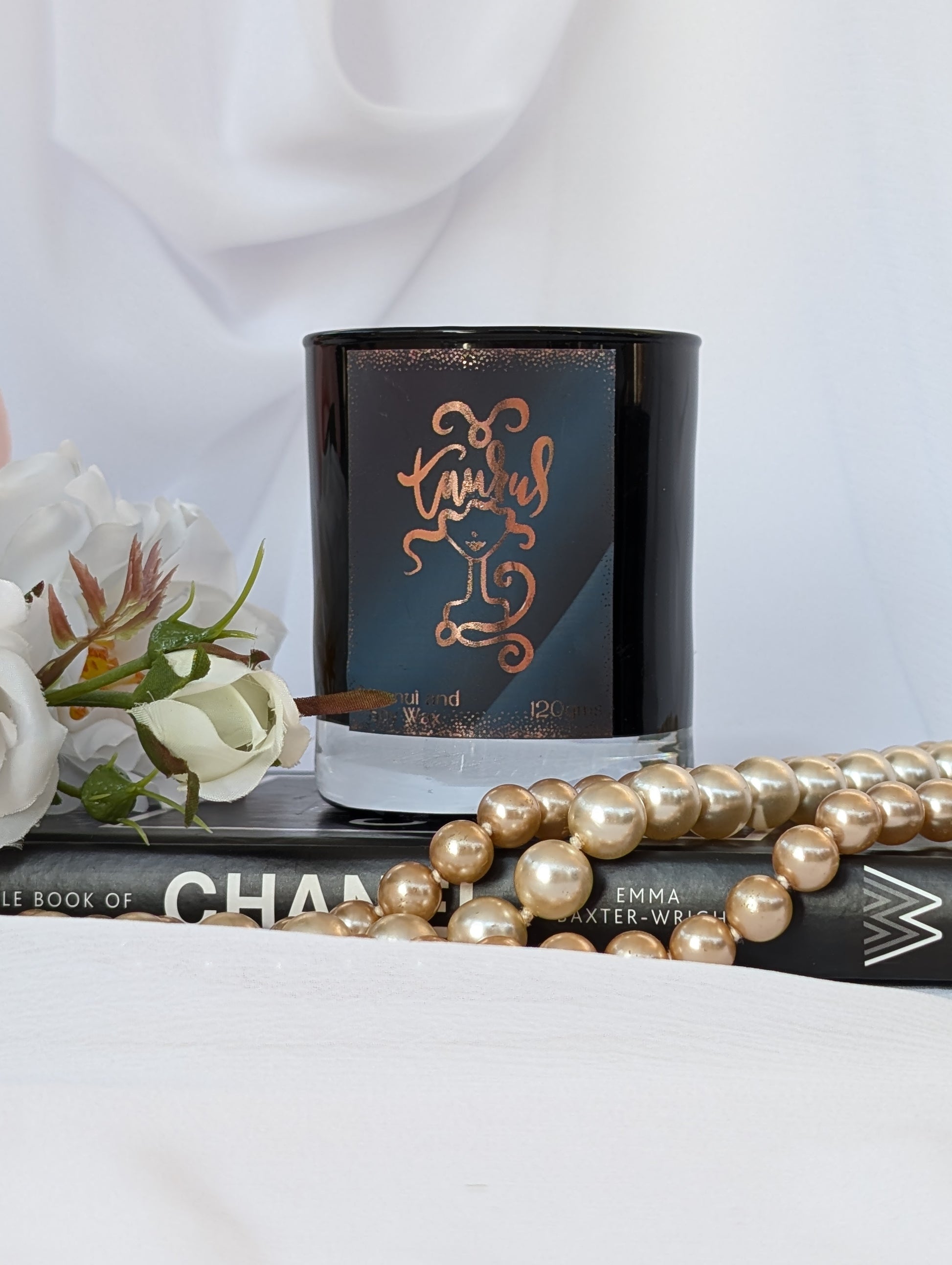Black and Gold Taurus candle - sits on a book, styled with white flowers and a strand of gold pearls against a soft white draped fabric background.