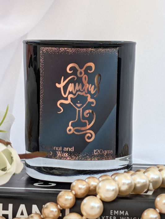 Taurus black and gold zodiac candle. with rose gold foil label.  The candle is styled on a book with pearl beads in the foreground against a white draped background.