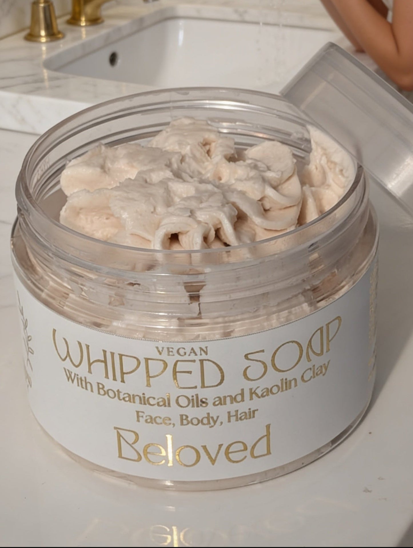 Woman applying whipped soap from a jar labeled 'Beloved' on a bathroom counter.