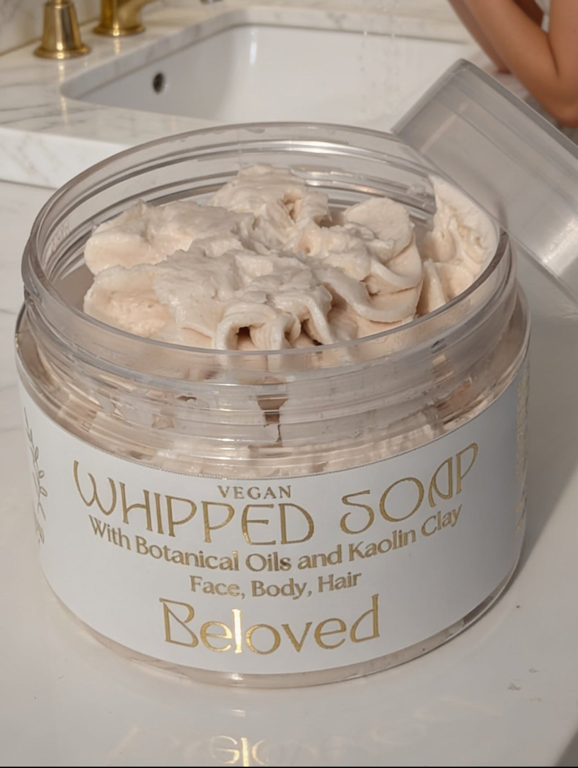 Woman applying whipped soap from a jar labeled 'Beloved' on a bathroom counter.