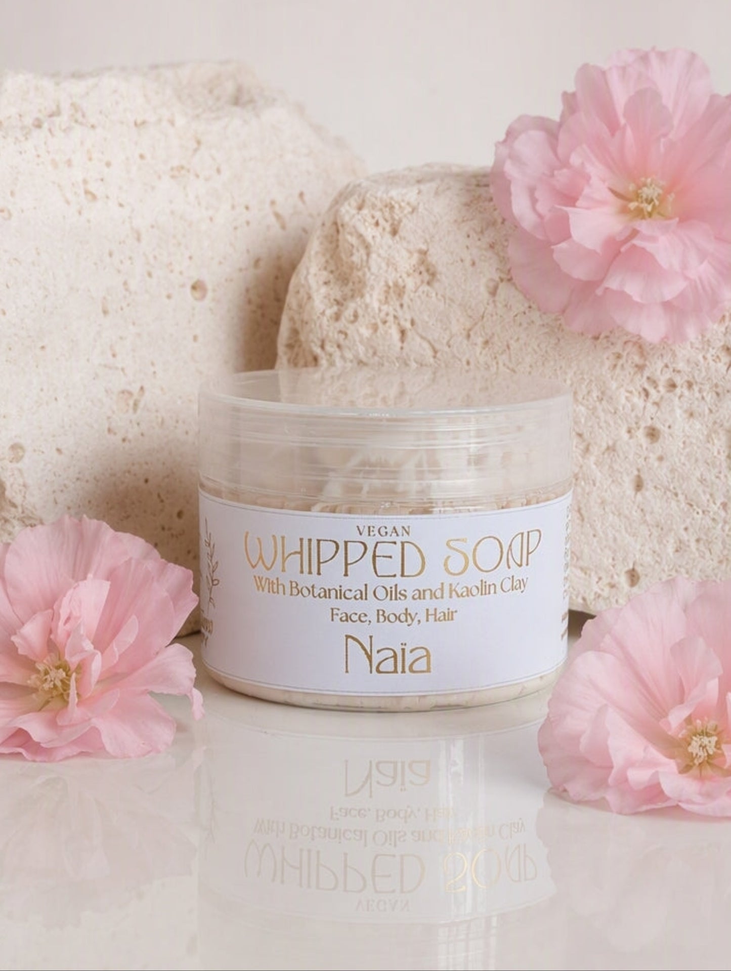 A jar of Naia Whipped Soap with botanical oils and hair care, styled with pink flowers and textured stone blocks on a reflective white surface.