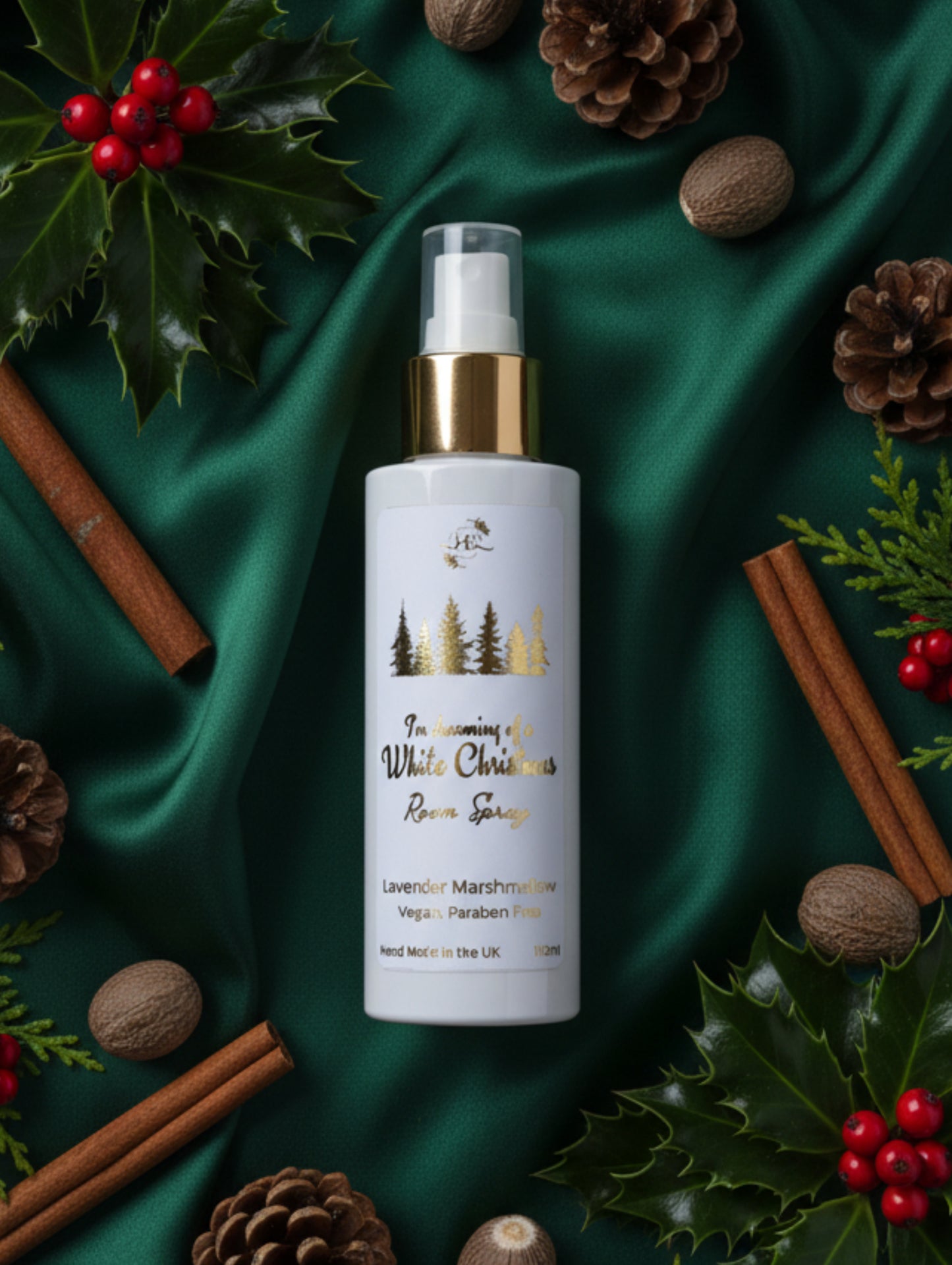A 100ml spray bottle of White Christmas room spray with gold cap, and gold lettering, set against a green satin draped cloth with pinecones, holly and nutmeg