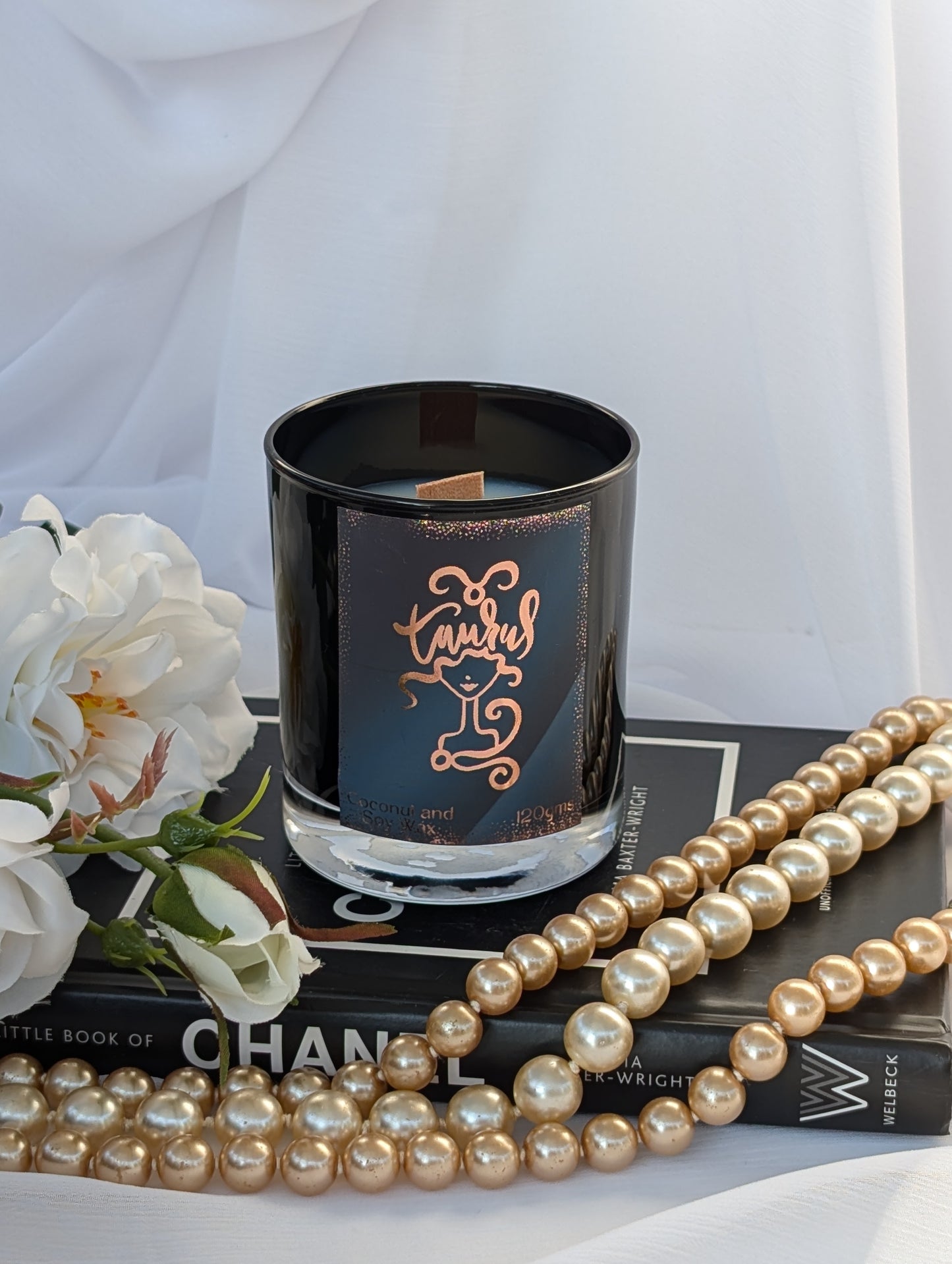 Black and Gold Taurus Zodiac Candle - styled on a black book with white flowers and a strand of gold faux pearls against a white draped fabric background.