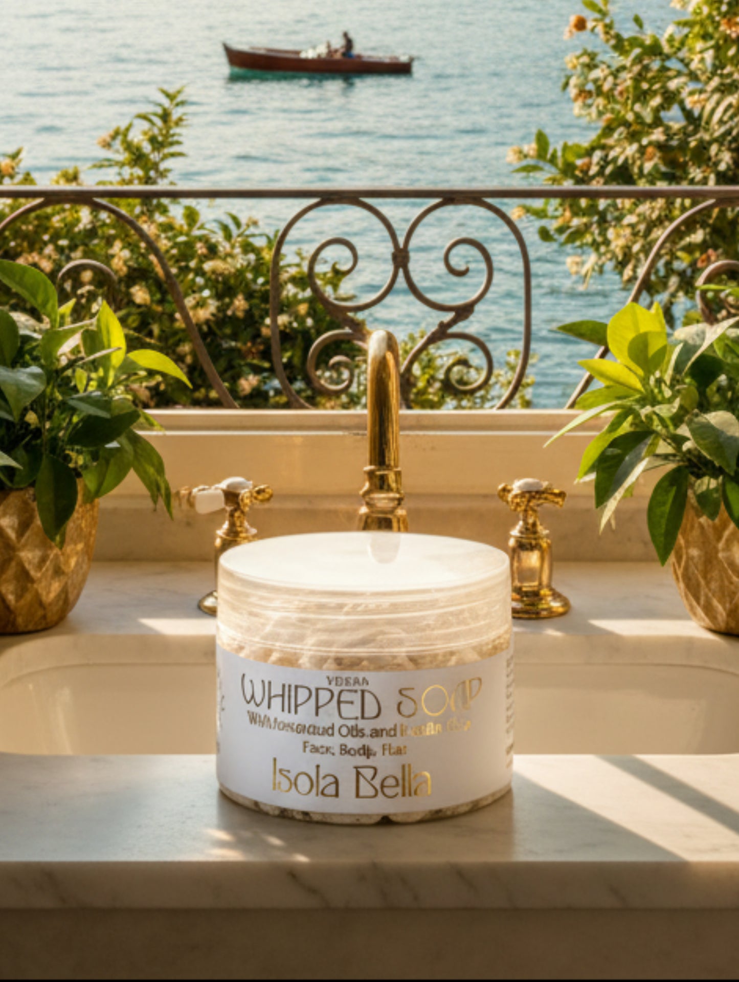A jar of Isola Bella Vegan Whipped Soap with botanical oils and kaolin clay, featuring a white label with gold lettering and a clear lid, set on a bathroom windowledge overlooking a lake